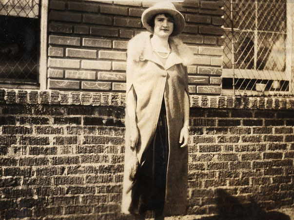 <lady posed by scool building's brick wall window guards wearing pearls hat fur color. cape style coat with slits for arms to protude through>