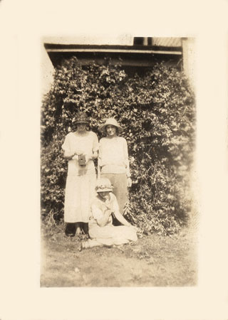 <three ladies posed in front of a vine. The person seated may be Johnie>