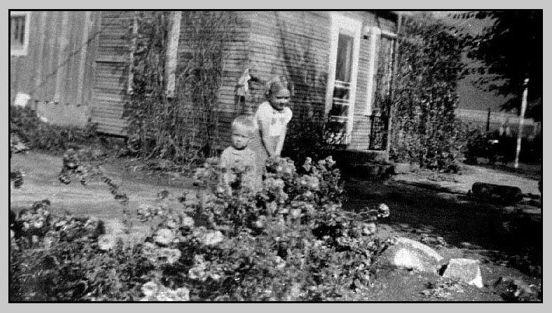 <Terry and Adrienne standing in yard of ranch house. Note rocks used to border the flowerbed>
