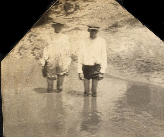 <Two boys wading in the river with their pants rolled up.>